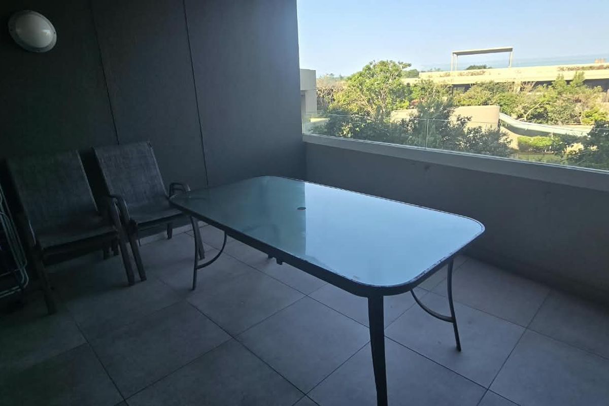 A covered balcony featuring tiled flooring, a glass balustrade, and a wall-mounted light fixture, offering an outdoor space for relaxation.