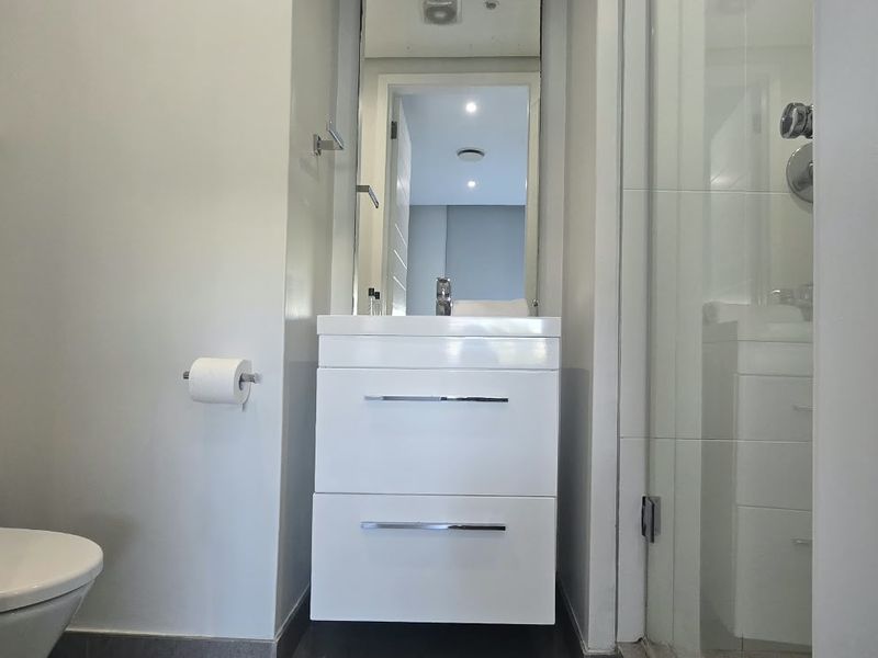 A modern, compact bathroom featuring a white vanity cabinet, wall-mounted toilet, and a glass-enclosed shower area with clean, contemporary finishes.