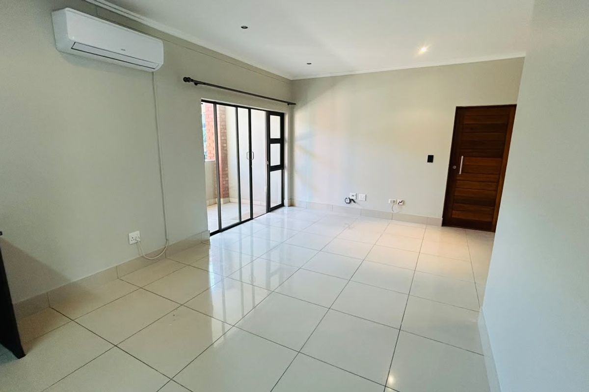 A bright, open-plan living area featuring neutral tiled flooring, a wall-mounted air conditioning unit, and sliding glass doors leading to an outdoor space.