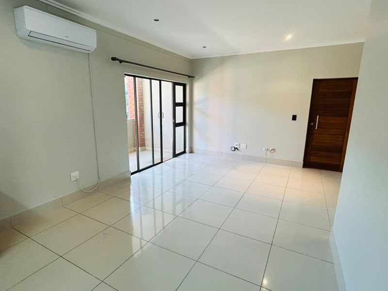 A bright, open-plan living area featuring neutral tiled flooring, a wall-mounted air conditioning unit, and sliding glass doors leading to an outdoor space.