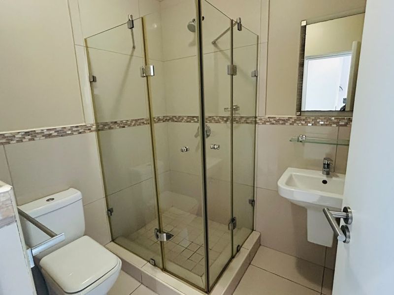 A clean, functional bathroom featuring a glass-enclosed shower, pedestal basin, and toilet with neutral wall and floor tiling.