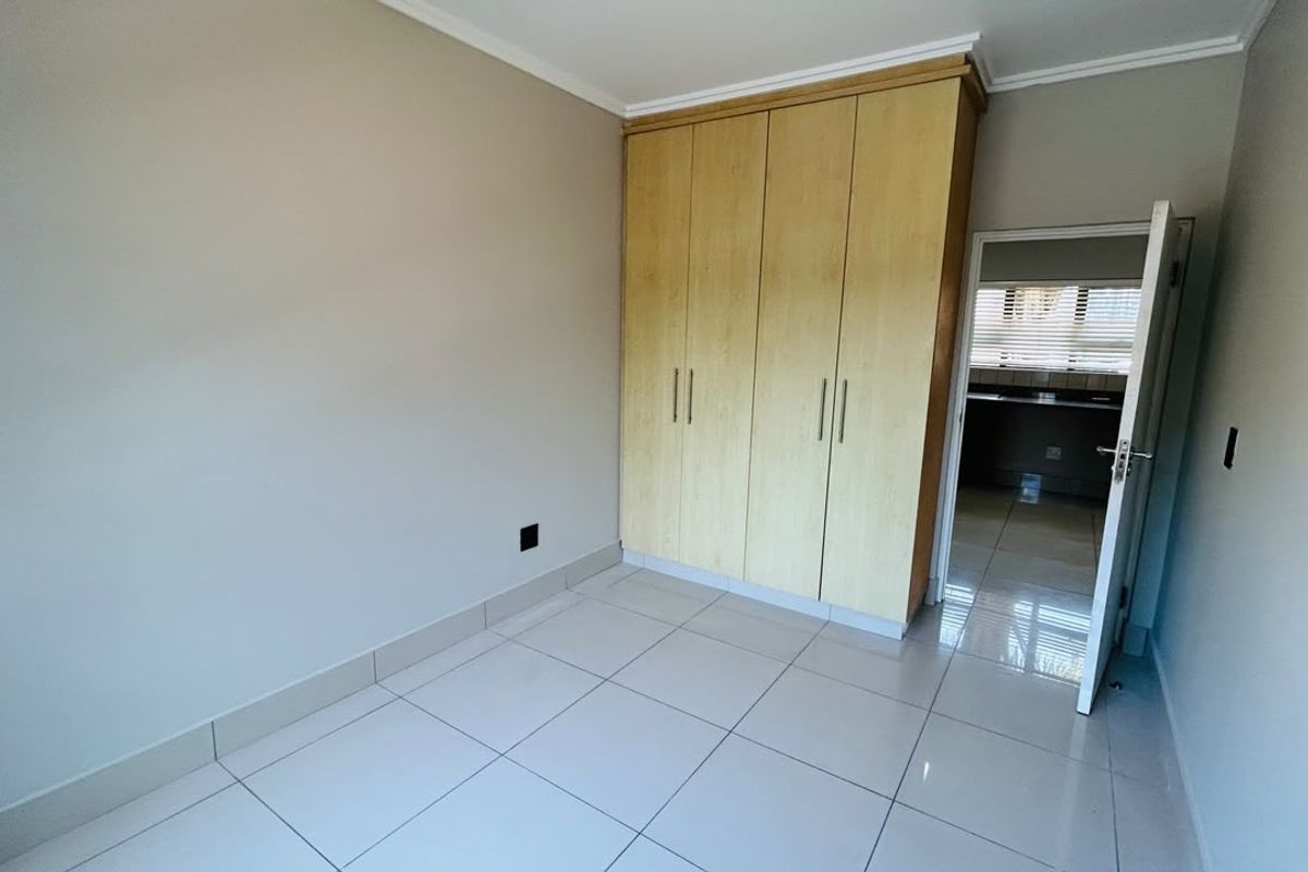 A clean, empty bedroom featuring neutral tiled flooring, built-in wooden cupboards, and a doorway leading to an adjacent room.