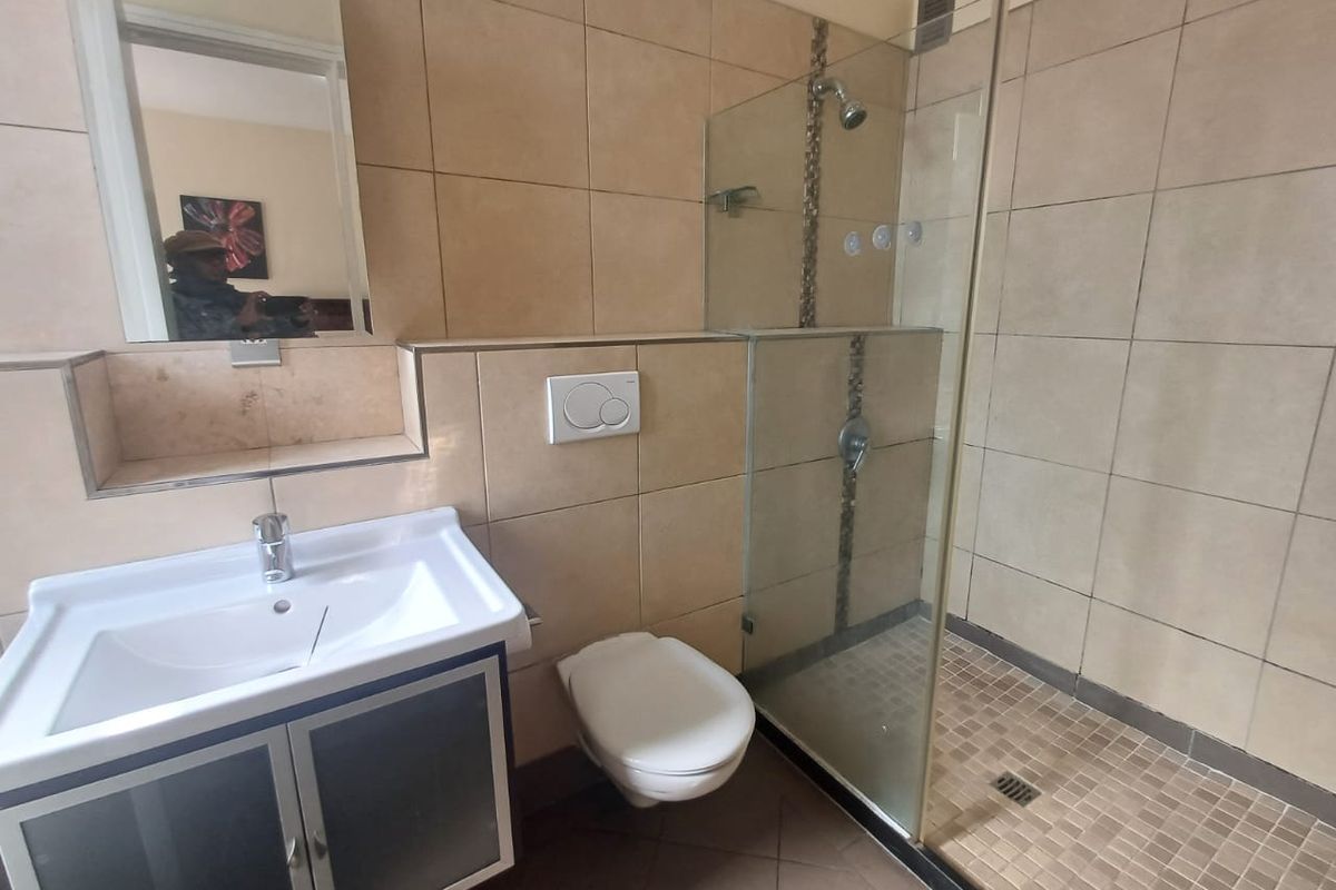 A functional bathroom featuring a glass-enclosed shower, wall-mounted toilet, and a vanity basin with a mirror, finished with neutral wall and floor tiles.