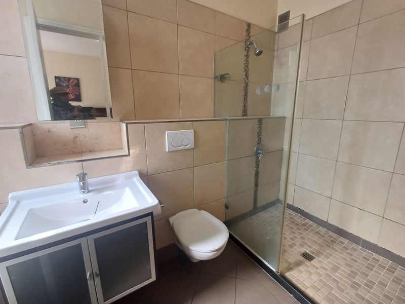 A functional bathroom featuring a glass-enclosed shower, wall-mounted toilet, and a vanity basin with a mirror, finished with neutral wall and floor tiles.