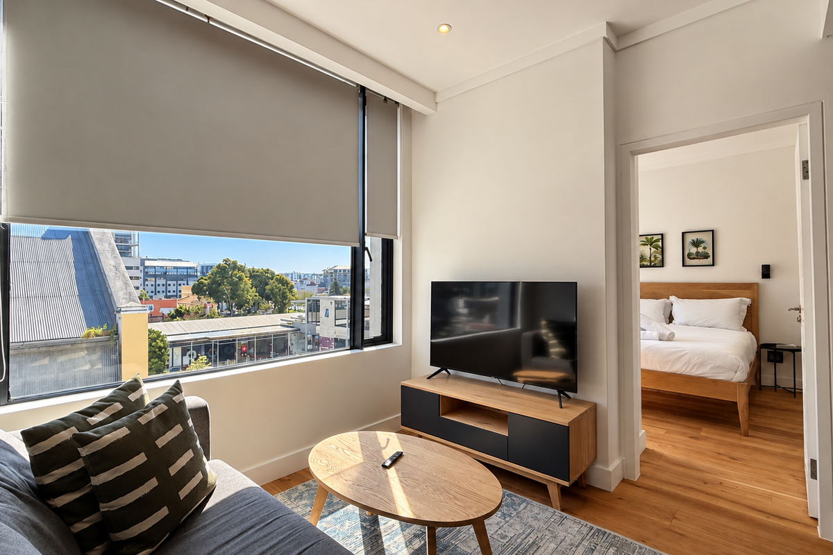 A bright living area featuring timber flooring, recessed lighting, and large windows with roller blinds, offering a view and access to the adjacent bedroom.