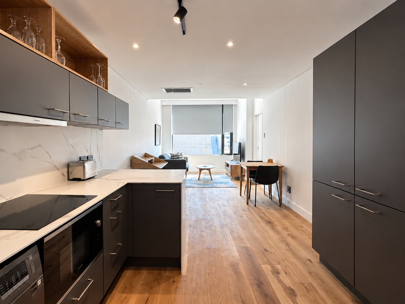 Modern open-plan apartment featuring a sleek kitchen with dark cabinetry, marble-look countertops, integrated appliances, and warm timber flooring throughout the living area.