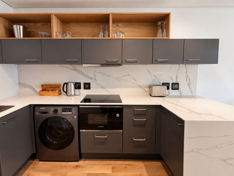 Modern kitchen featuring dark grey cabinetry, marble-look stone countertops and backsplash, integrated washing machine, built-in oven, and sleek open shelving.
