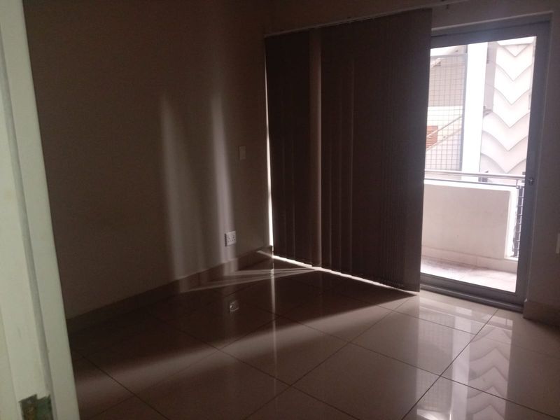 An empty bedroom featuring tiled flooring and a sliding glass door leading to a balcony, providing natural light through vertical blinds.