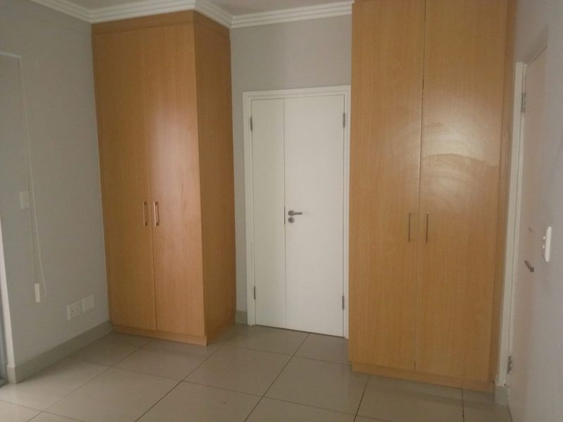 A clean, empty bedroom featuring light wood-finish built-in cupboards, neutral tiled flooring, and white walls with decorative cornice detailing.