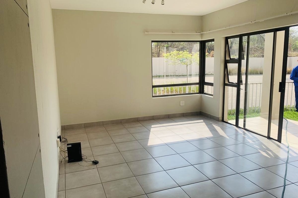 A bright, empty living room featuring neutral tiled flooring, large corner windows, and a sliding glass door providing access to the outdoor area.
