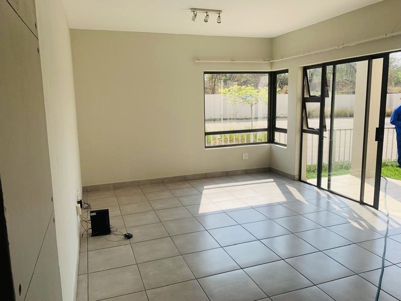 A bright, empty living room featuring neutral tiled flooring, large corner windows, and a sliding glass door providing access to the outdoor area.