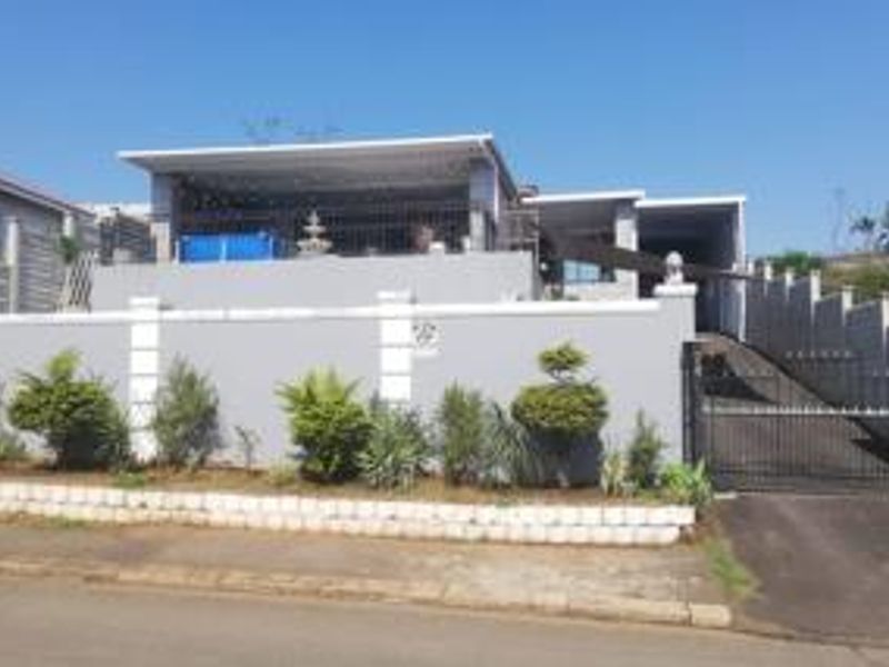 Front elevation of a residential property featuring a boundary wall, paved driveway, metal gate, and an elevated covered patio area.