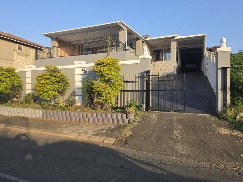 Front elevation of a residential property featuring a paved driveway, secure wrought iron gate, raised patio area, and a covered carport.