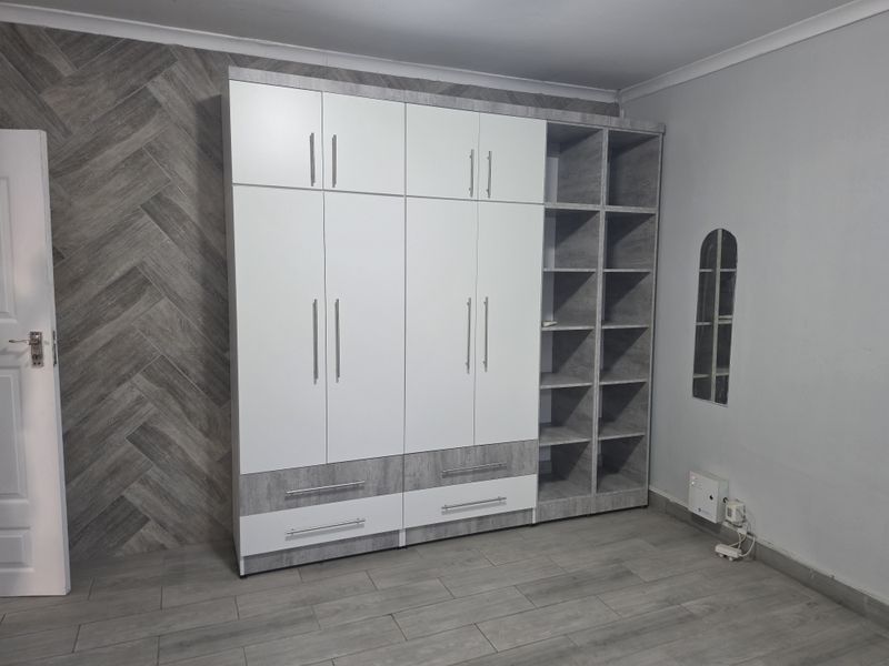A modern bedroom featuring extensive built-in cabinetry with white and grey wood-grain finishes, complemented by matching laminate flooring and a decorative feature wall.