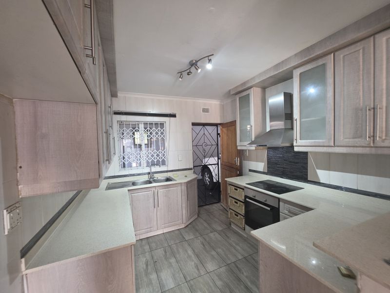 A functional kitchen featuring light wood-finish cabinetry, stone countertops, integrated oven and hob, and tiled flooring with a security gate leading outdoors.