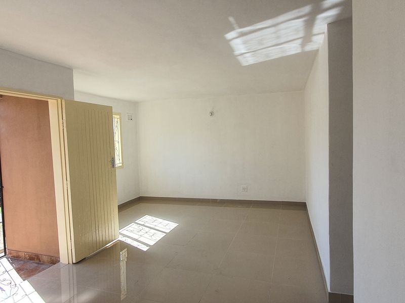 An empty, sunlit living area featuring neutral tiled flooring, security bars on windows, and direct access to an outdoor space.