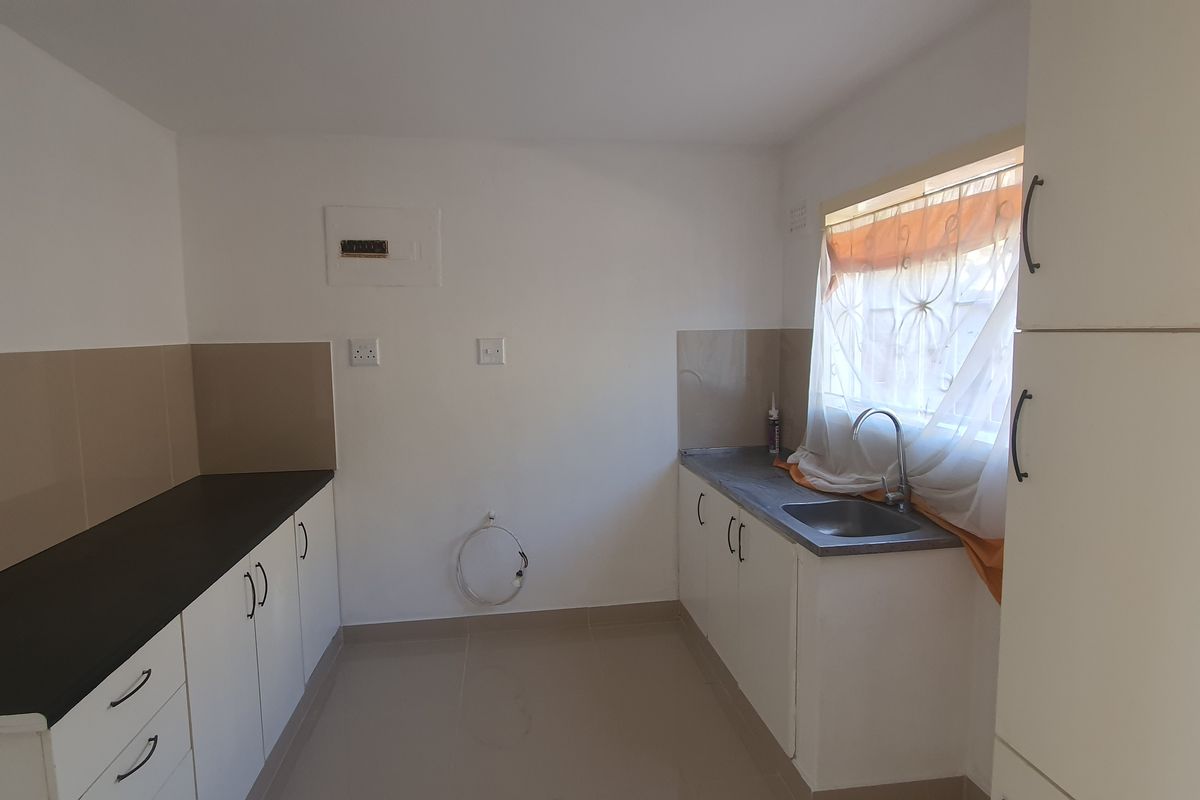 A clean, empty kitchen featuring white built-in cabinetry, dark countertops, a stainless steel sink, and neutral tiled flooring.