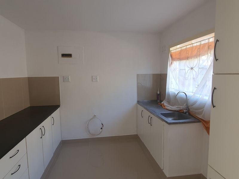 A clean, empty kitchen featuring white built-in cabinetry, dark countertops, a stainless steel sink, and neutral tiled flooring.