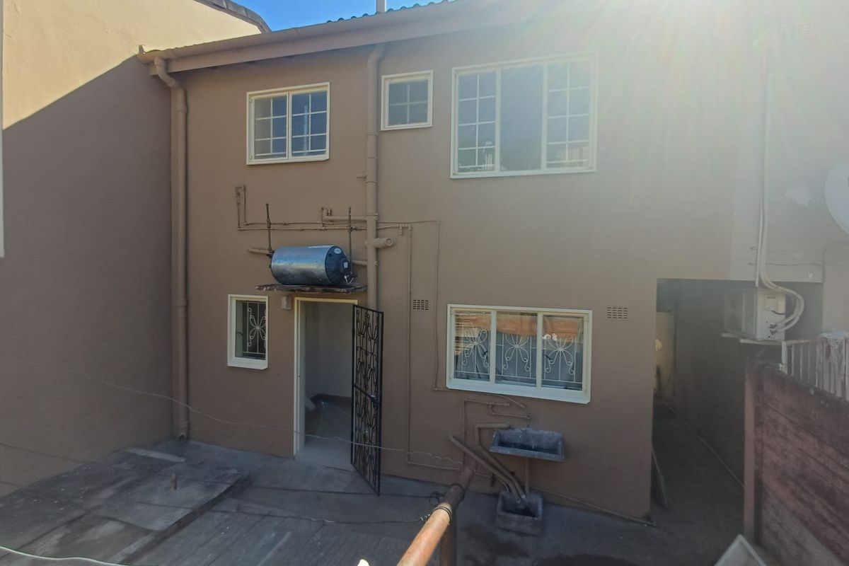 Rear exterior view of the townhouse featuring a paved courtyard, external geyser, and fixed laundry basin with security gate access.