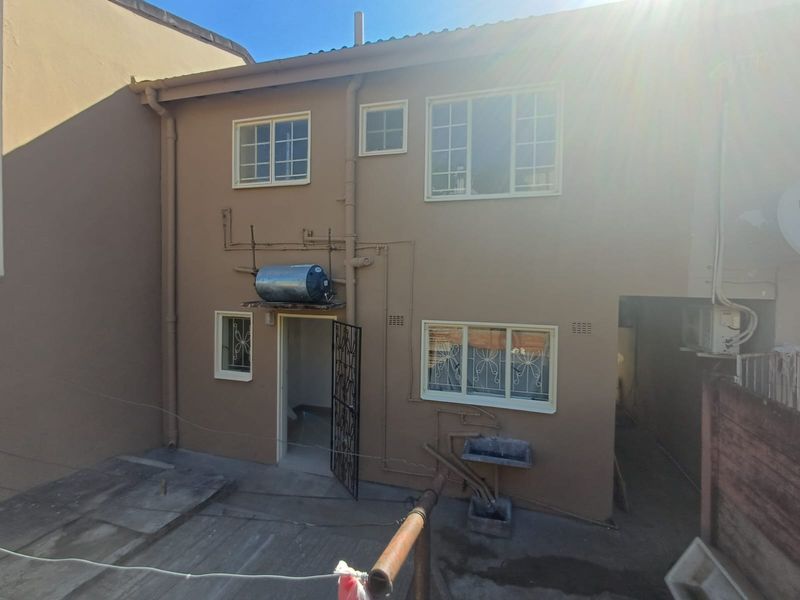 Rear exterior view of the townhouse featuring a paved courtyard, external geyser, and fixed laundry basin with security gate access.