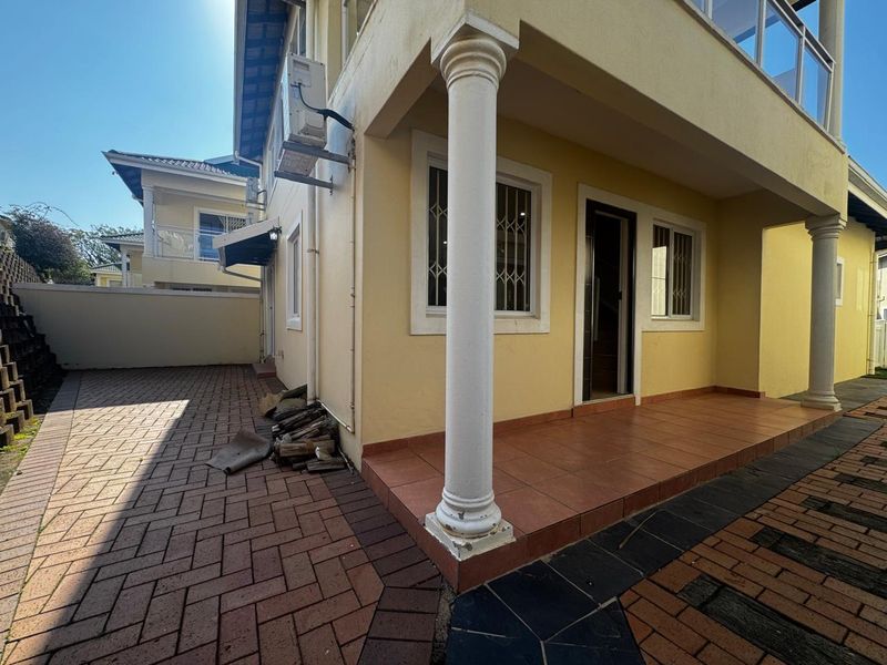 Rear exterior view of the house featuring a paved driveway, a covered tiled patio with decorative columns, and security bars on the windows.
