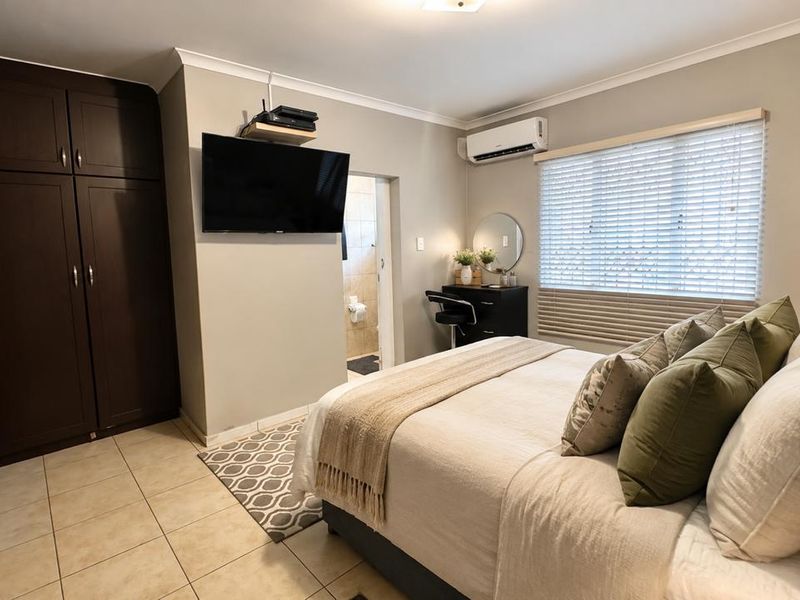 A well-maintained bedroom featuring built-in cupboards, tiled flooring, an air conditioning unit, and venetian blinds, with an en-suite bathroom access.