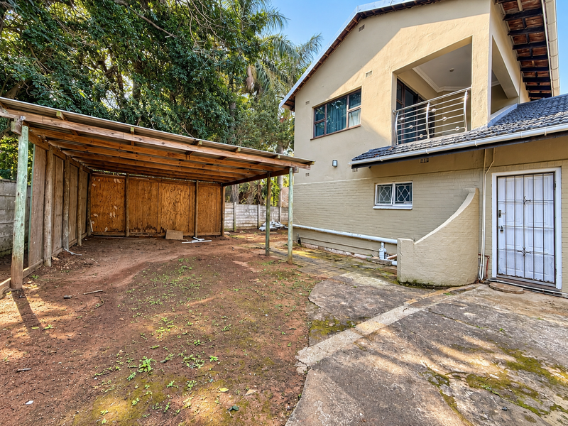 Rear exterior view featuring a timber carport, paved patio area, and a balcony with stainless steel railing attached to the main house structure.