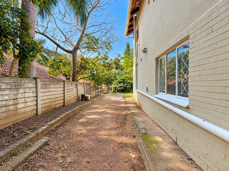 A side exterior view of the property featuring a painted brick wall, leaded glass windows, and a precast concrete boundary wall along a dirt path.