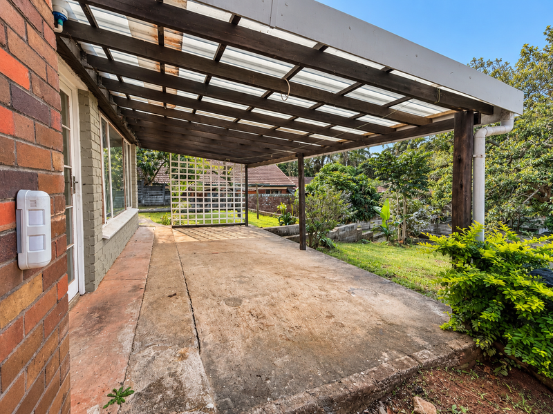 A covered outdoor patio area featuring a timber pergola structure and concrete flooring, overlooking the rear garden space.