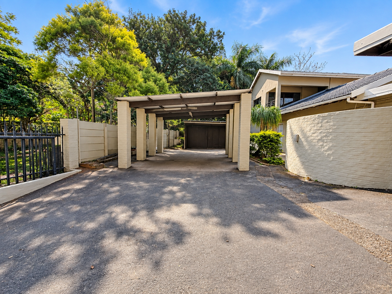A paved driveway leading to a covered carport and a separate garage, featuring brick boundary walls and established garden surroundings.