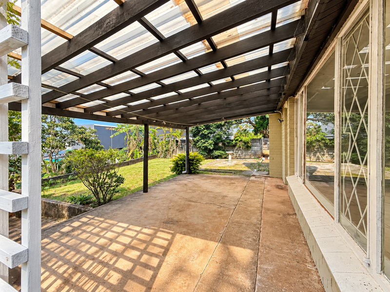 A covered outdoor patio area featuring a timber pergola with translucent roofing, concrete flooring, and direct access to the garden through large sliding glass doors.