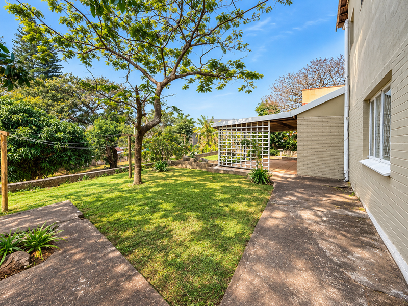 Rear exterior view featuring a covered patio area, concrete paving, and a well-established garden with mature trees.