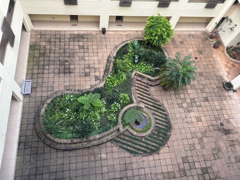 An elevated view of a central apartment complex courtyard featuring paved flooring, a landscaped garden bed with a small water feature, and surrounding walkways.