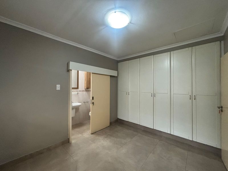 A spacious bedroom featuring neutral tiled flooring, extensive built-in cupboards, and direct access to an en-suite bathroom.