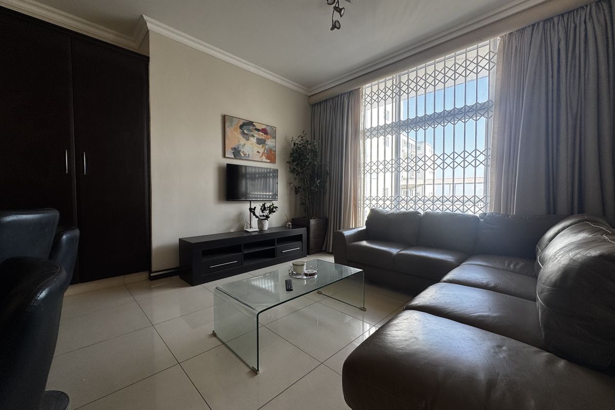 A well-maintained living room featuring large windows with security gates, tiled flooring, and a built-in cupboard for storage.