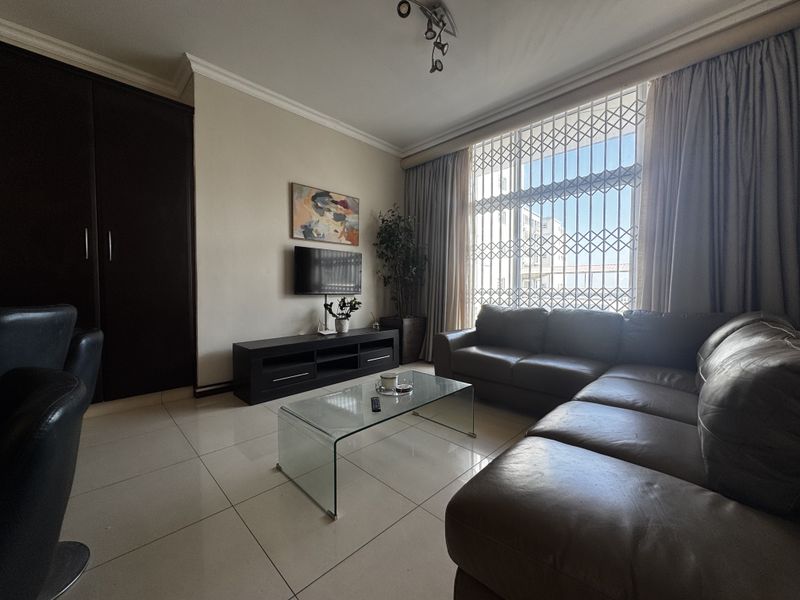A well-maintained living room featuring large windows with security gates, tiled flooring, and a built-in cupboard for storage.