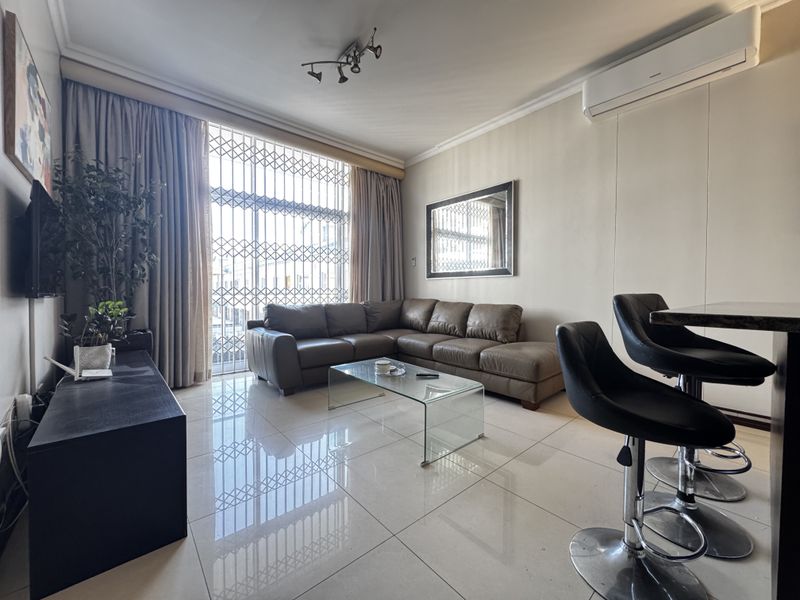 Modern open-plan living area featuring polished porcelain floor tiles, a wall-mounted air conditioning unit, and a breakfast bar, with large windows and security gate.