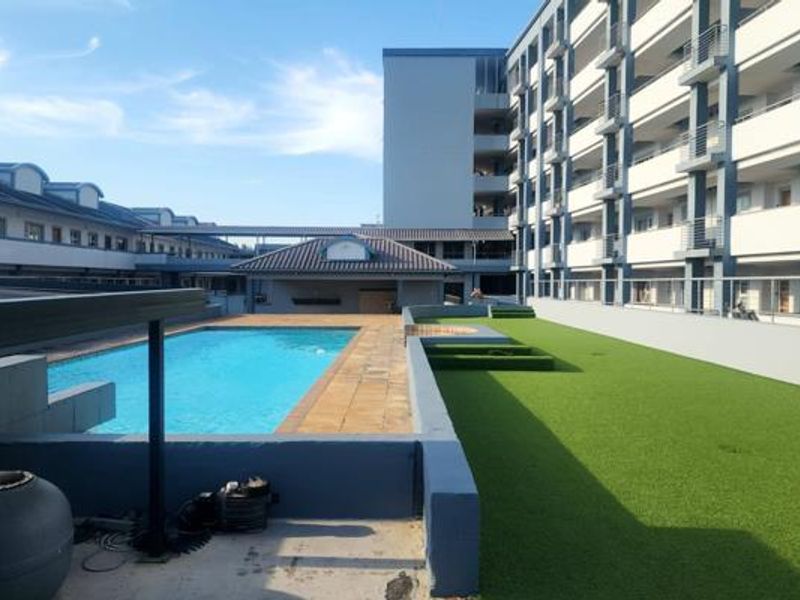 A communal pool area featuring a swimming pool, paved decking, and an artificial grass lawn, set within a multi-story apartment complex.