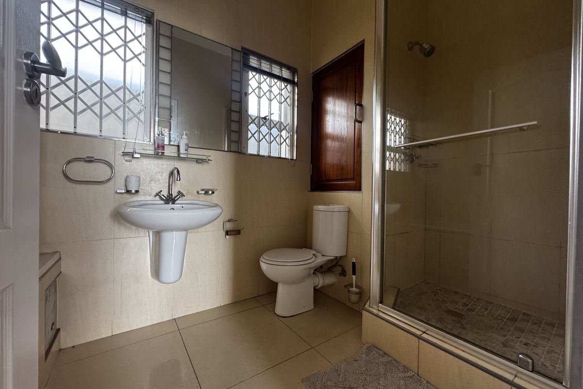 A functional bathroom featuring a pedestal washbasin, toilet, and a glass-enclosed shower with tiled finishes and security bars on the windows.