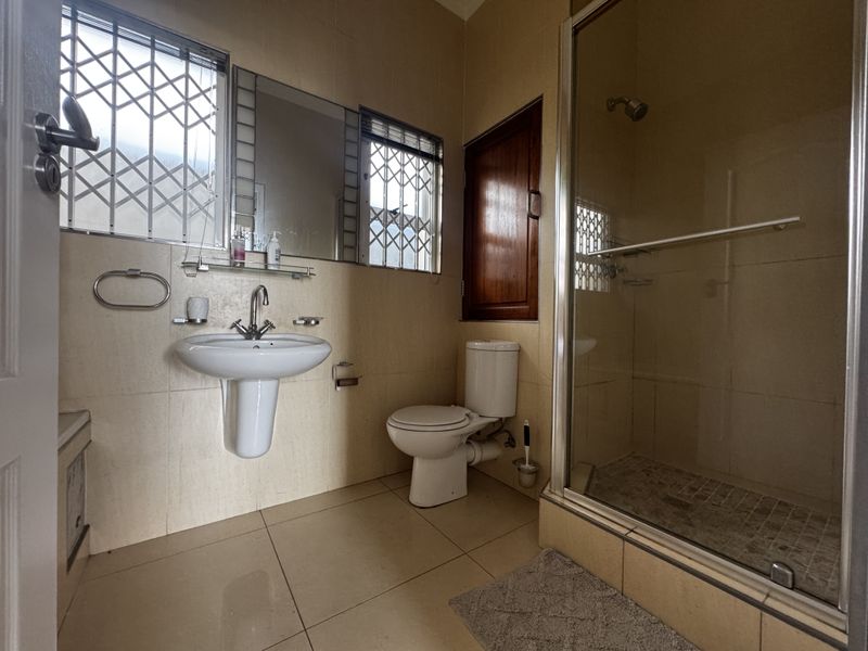 A functional bathroom featuring a pedestal washbasin, toilet, and a glass-enclosed shower with tiled finishes and security bars on the windows.