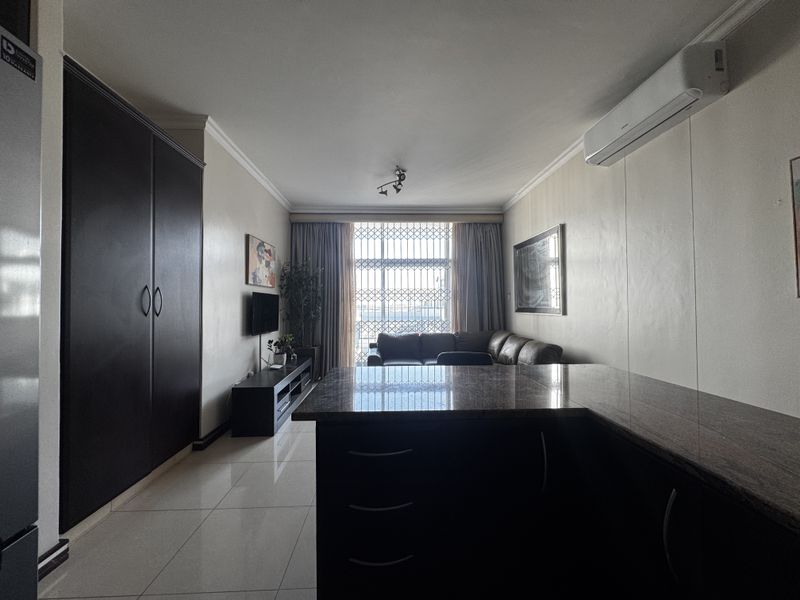 Open-plan living area featuring tiled floors, a granite-topped kitchen counter, and a wall-mounted air conditioning unit, leading to a window with security screening.