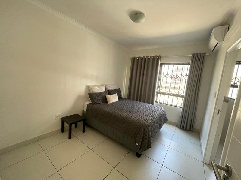 A well-maintained bedroom featuring tiled flooring, an integrated air conditioning unit, and a window fitted with a security gate.