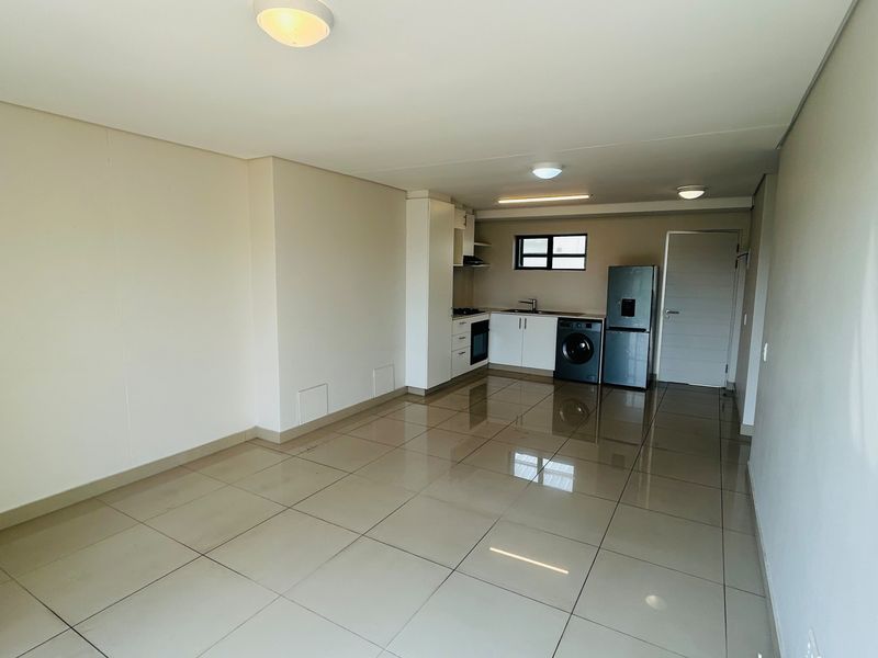 An open-plan living and kitchen area featuring neutral tiled flooring, modern built-in cabinetry, and integrated kitchen appliances in a clean, vacant space.