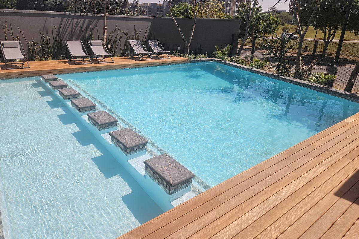 A well-maintained swimming pool area featuring timber decking, a stepped entry, and a secure boundary wall in a modern apartment complex setting.