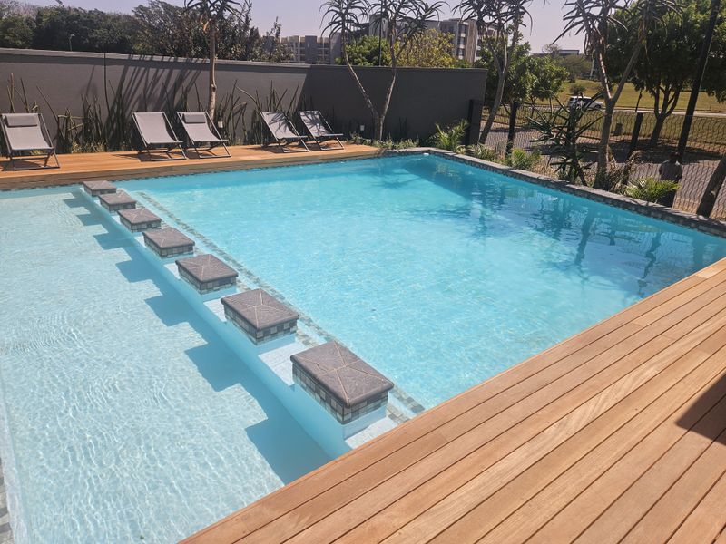 A well-maintained swimming pool area featuring timber decking, a stepped entry, and a secure boundary wall in a modern apartment complex setting.