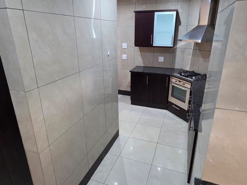 A compact, modern kitchen featuring dark cabinetry, granite countertops, a gas hob, built-in oven, and neutral floor-to-ceiling wall tiles.