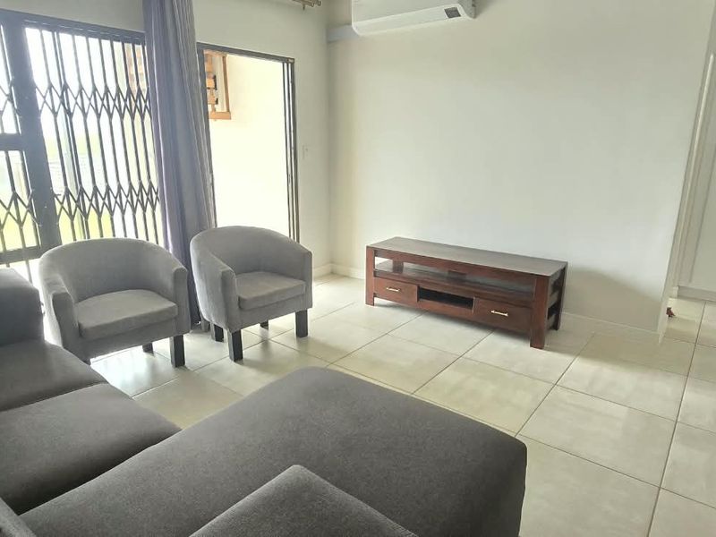 A bright living room featuring tiled flooring, a wall-mounted air conditioning unit, and a sliding door with a security gate leading to the exterior.