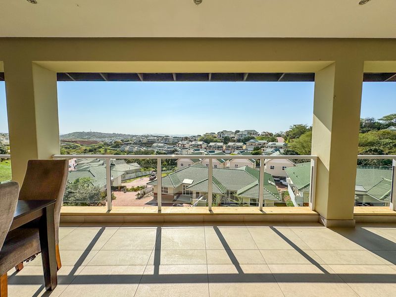 A spacious covered balcony featuring tiled flooring and a modern glass balustrade, offering expansive elevated views over the surrounding residential estate.