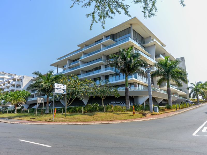 A modern multi-story apartment building featuring tiered balconies with glass balustrades, situated on a corner lot with landscaped surroundings.