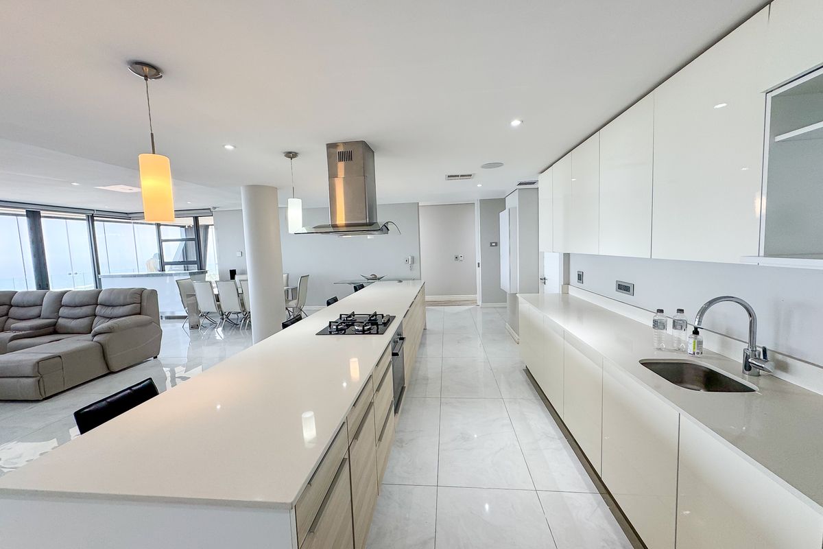 Modern open-plan kitchen featuring a long island with gas cooktop, sleek gloss cabinetry, stone countertops, and integrated appliances, flowing into the living and dining area.